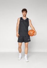 Camiseta sin mangas de baloncesto negra con ribetes blancos, combinada con pantalones cortos negros que presentan el logo "NETS". El modelo sostiene un balón de baloncesto y lleva zapatillas blancas.