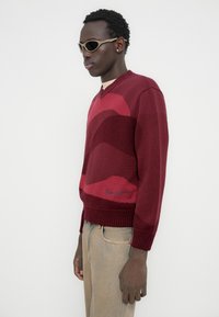 Μπορντό πλεκτό πουλόβερ με κυματιστά χρωματικά σχέδια, σχεδιασμό με λαιμόκοψη V, ριμπ μανσέτες και λεπτομέρειες στο τελείωμα. Συνδυασμένο με μπεζ παντελόνια και γυαλιά ηλίου.