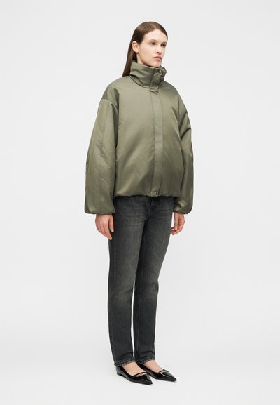 Γυναίκα που στέκεται φορώντας μια φαρδιά μπουφάν φουσκωτή σε ελιά πράσινο, μαύρα τζιν και μαύρα παπούτσια με μυτερή μύτη, μπροστά σε απλό φόντο.
