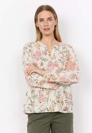 Frau mit glatten blonden Haaren, die eine cremefarbene Langarmbluse mit rosa Blumenmustern und olivgrüne Hose trägt, die Arme verschränkt.