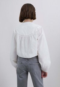 Blusa blanca de manga larga con mangas abullonadas, con detalles fruncidos en la parte trasera. Combinada con jeans ajustados grises.
