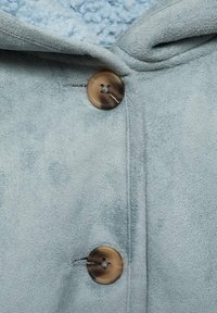 Detail světle modrého fleecového kabátu s měkkým, texturovaným povrchem, kapucí a velkým kulatým hnědým knoflíkem se čtyřmi dírkami.