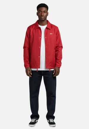 Giovane uomo in piedi, che indossa una giacca rossa con bottoni sopra una t-shirt bianca, jeans scuri e sneakers nere con lacci bianchi.