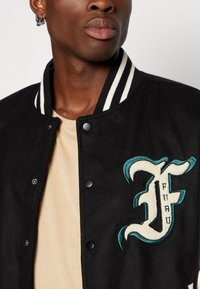 Black suede jacket with a striped collar, snap buttons, and an embroidered letter "F" in teal and cream on the chest. Cream shirt underneath.