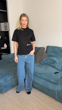 Femeie cu păr blond, purtând un tricou negru și blugi albaștri largi, stă lângă o canapea albastră cu perne într-un living.