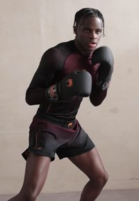 Αθλητικός άνδρας σε εφαρμοστό μαύρο και βουργουνδί μπλουζάκι και μαύρες βερμούδες, φορώντας μαύρα γάντια πυγμαχίας με πορτοκαλί λεπτομέρειες, προετοιμάζεται για πάλη.