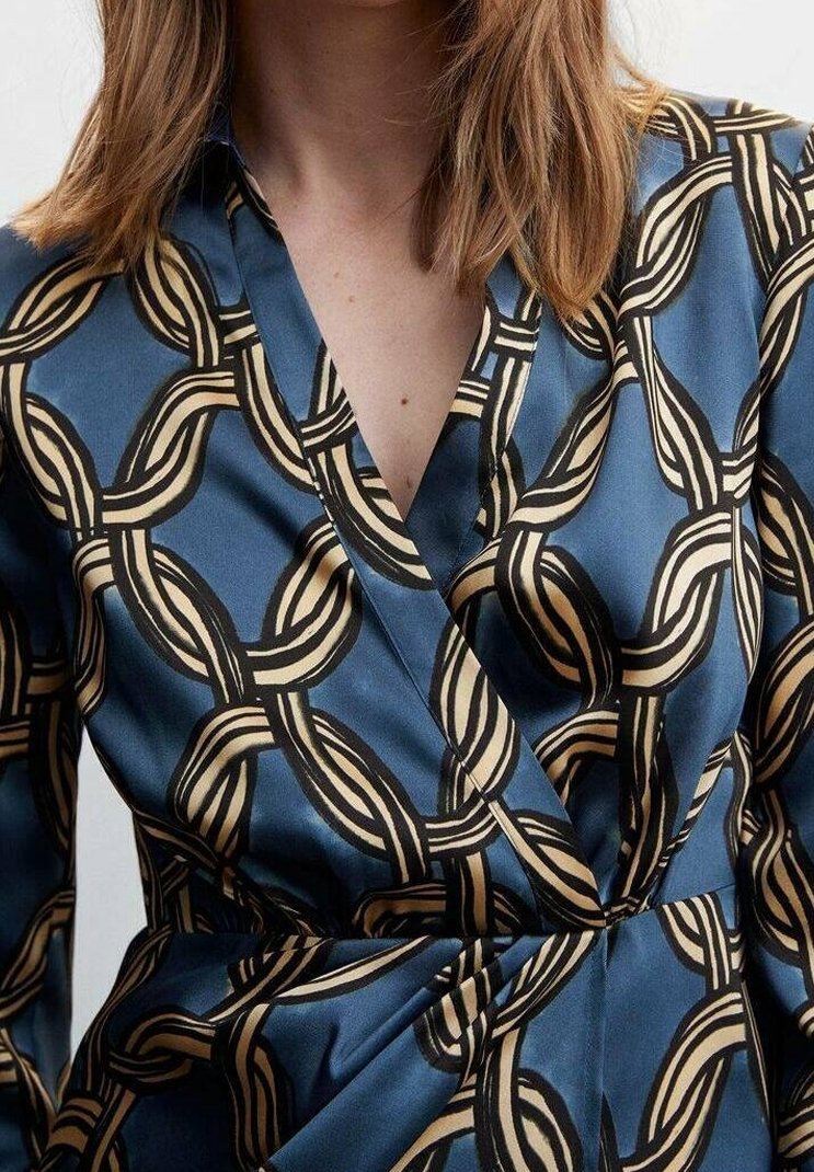 Femme portant une robe portefeuille bleue avec un motif de chaînes beige et noir entrelacées, dévoilant le décolleté et la taille.