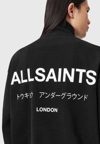 Melna mikstere ar izteiksmīgu baltu tekstu, kurā rakstīts "ALLSAINTS", un japāņu rakstzīmēm, zem kurām ir norādīts "LONDON". Augsta apkakles dizains.