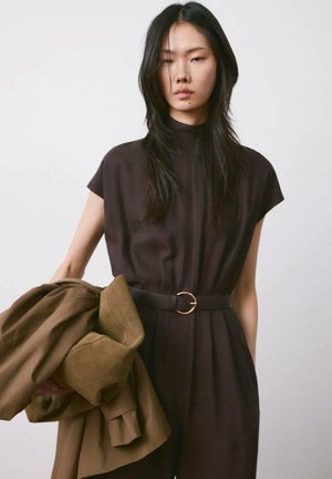 Mujer con cabello largo y negro, vestida con un vestido oscuro con cinturón, sosteniendo un abrigo marrón doblado sobre un fondo blanco liso.