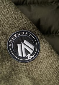 Tessuto a maglia verde oliva con un patch ricamato rotondo bianco e nero con la scritta "SUPERDRY" e dettagli di design geometrico.