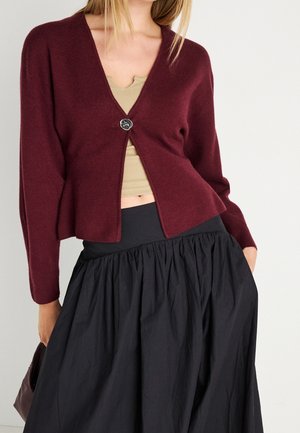 Femme portant un cardigan bordeaux à un bouton sur un haut beige et une jupe noire froncée taille haute, main dans la poche de la jupe.