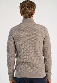 Beige tröja med diamantmönster, ribbad krage och muddar, strukturerad tyg. Bakifrån visas en åtsittande design och sidologotyp.