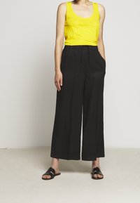 Henkilö, jolla on päällä keltainen hihaton paita, mustat leveät housut ja mustat avonaiset liukujalkineet, seisoo kädet taskuissa tasaisella lattiapinnalla.