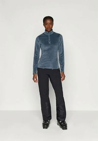 Γυναίκα που φοράει μπλε φούτερ με φερμουάρ, μαύρο παντελόνι και μαύρες μπότες χιονιού, στέκεται μπροστά σε λευκό φόντο.