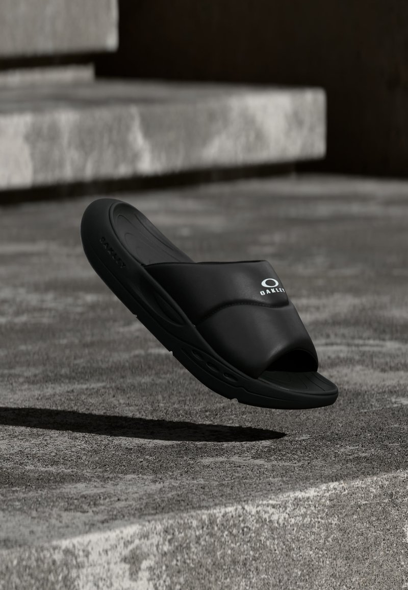 Μαύρο σανδάλι Oakley slide αιωρούμενο πάνω από υφή σκαλοπατιών από σκυρόδεμα υπό απαλό φωτισμό, εμφανίζοντας πλευρική και κορυφαία όψη με ορατό λογότυπο.