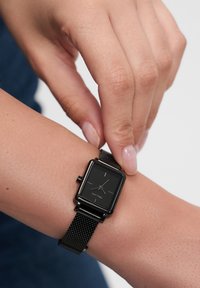 Montre carrée noire avec un bracelet en maille. Présente un cadran noir simple avec des aiguilles argentées et de petits index. Fixée à un poignet.