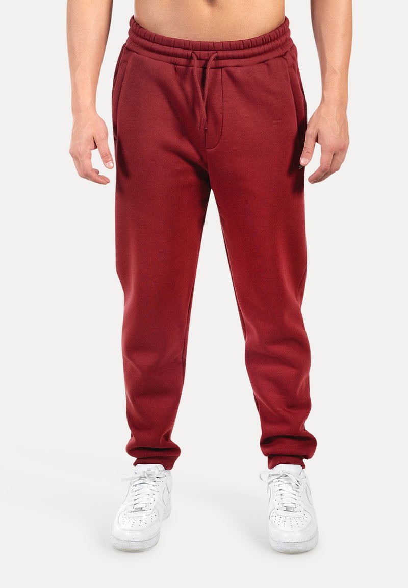 Muž v burgundských teplákách s šňůrkou v pase a bílých teniskách stojící na jednobarevném pozadí.