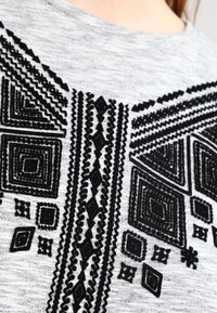 T-shirt grigia con intricato ricamo geometrico nero che presenta quadrati, diamanti e motivi a zigzag sulla zona del petto.