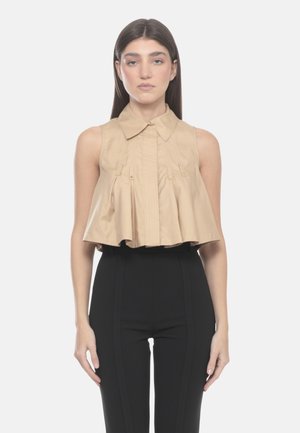 Donna che indossa una camicetta beige smanicata corta con colletto e pieghe, abbinata a pantaloni neri aderenti a vita alta, in piedi su sfondo bianco.