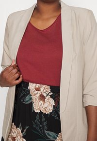 Blazer beige chiaro sopra una maglia bordeaux, abbinati a una gonna nera con grandi fiori crema e dettagli verdi.