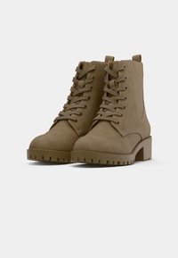 Paire de bottines en daim beige avec talons bas épais, lacets sur le devant et semelles épaisses à crampons sur fond blanc.