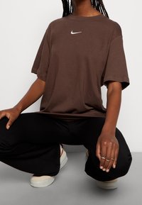 Hnědé oversized tričko s bílým logem Nike na hrudi, spárované s černými kalhotami s širokými nohavicemi a krémovými botami.