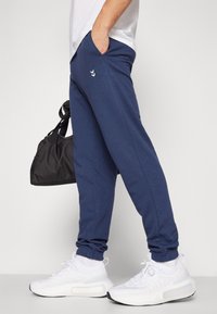 Marineblaue Jogginghosen aus weichem Stoff, mit Seitentaschen und gesticktem Logo. Kombiniert mit weißen Sneakers und einer schwarzen Tasche.