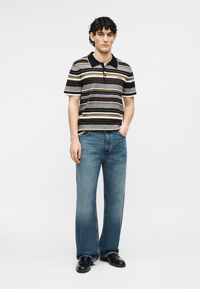 Νέος άνδρας με σγουρά μαλλιά, που φοράει μπλούζα πόλο με ρίγες και μαύρο γιακά, μπλε τζιν και μαύρα παπούτσια, στέκεται με το ένα χέρι στην τσέπη.