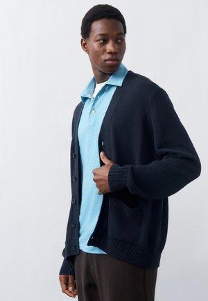 Νεαρός άνδρας που φοράει ένα ανοιχτό γαλάζιο πόλο πουκάμισο κάτω από ένα σκούρο μπλε ζακέτα με κουμπιά, κοιτάζει προς τα αριστερά του μπροστά σε ένα απλό φόντο.