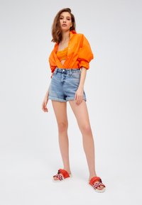 Pomarańczowa, oversize'owa koszula, prążkowany pomarańczowy crop top, jasnoniebieskie szorty z wysokim stanem oraz sandały w czerwono-białe wzory.