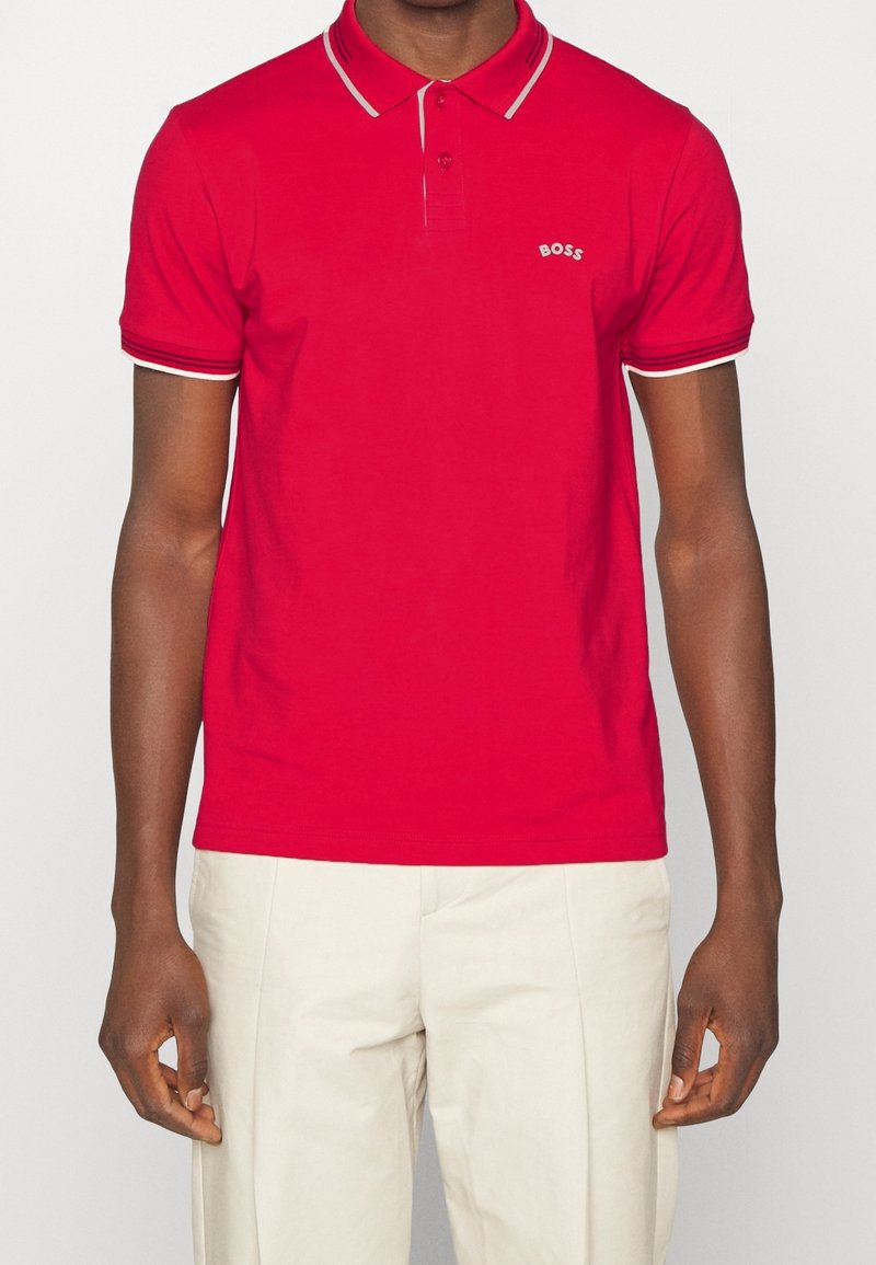 Röd bomullspikétröja med korta ärmar, en placket med två knappar och kontrasterande vita ränder på kragen och ärmarna. Litet logotyp på bröstet.