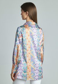 Camicia di seta con un motivo geometrico colorato in tonalità pastello, con colletto e maniche a sbuffo, mostrata di lato.