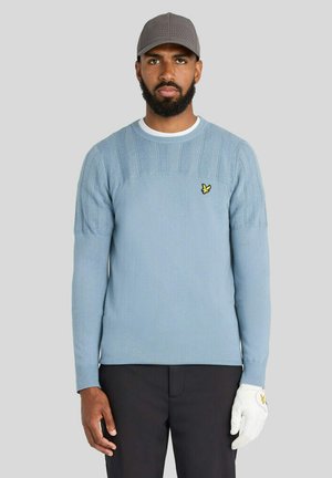 Mand iført en lyseblå sweater med en gul fuglelogo, sorte bukser, grå kasket og hvid golfhandske på højre hånd står mod en ensfarvet baggrund.