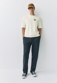 Valkoisen ja kellertävän sävyn puuvillainen t-paita, jossa pieni painatus rinnassa, yhdistetty tummanvihreisiin jogger-housuihin ja mustiin kangastennareihin. Ruskea samettimyssy.