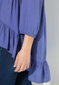 Blusa viola con orlo arricciato e maniche a tre quarti, realizzata in tessuto morbido. Mano appoggiata in basso vicino all'orlo.