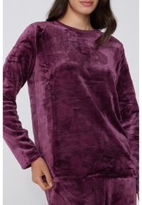 Top púrpura de terciopelo con mangas largas, escote redondo y textura suave. Presenta mangas con puños y un dobladillo recto para un ajuste relajado.