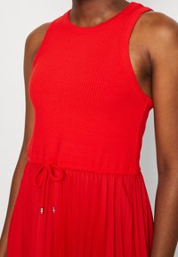 Punainen ribbikleid, jossa on olkaimeton muotoilu, vyötäröä korostava henkäilevä laskostettu hame ja narulukot metallipäillä.