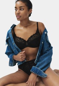 Vrouw die een zwarte kanten beha en ondergoedset draagt, gedeeltelijk bedekt door een open blauwe spijkerjasje, kijkt opzij, zittend op de grond.