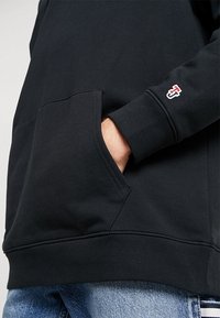 Sudadera negra con bolsillo frontal, puños y dobladillo acanalados. Presenta un pequeño logo en la manga. Textura suave. Combinada con vaqueros azul claro.