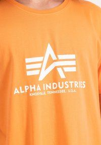 Oranges Baumwoll-T-Shirt mit einem auffälligen weißen Logo und dem Text "ALPHA INDUSTRIES" sowie geografischen Details darunter.