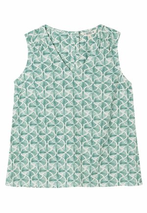 Junge Frau mit langen braunen Haaren, die ein ärmelloses grünes gemustertes Top und weiße Hosen trägt, steht vor einem hellen Hintergrund.