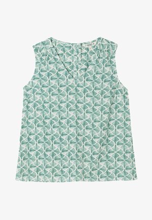 Junge Frau mit langen braunen Haaren, die ein ärmelloses grünes gemustertes Top und weiße Hosen trägt, steht vor einem hellen Hintergrund.