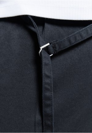 Close-up van een zwarte stoffen riem die door een kleine zilveren metalen lus is geregen, tegen een witte, textuurrijke achtergrond.