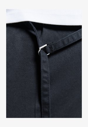 Close-up van een zwarte stoffen riem die door een kleine zilveren metalen lus is geregen, tegen een witte, textuurrijke achtergrond.