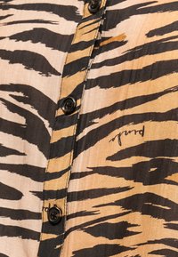 Κοντινό πλάνο πουκάμισου με μαύρα κουμπιά και ρίγες ζέβρας σε τόνους καφέ, κρεμ και μαύρου, με μικρή τυπωμένη λεπτομέρεια υπογραφής.