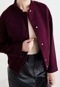 Bordeaux wollen jas met een ronde kraag, drukknoopjes en een boxy pasvorm, gedragen over een beige top en gecombineerd met een zwarte leren rok.