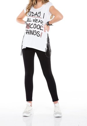 Kvinde iført en hvid ærmeløs top med teksten ""Today I will wear cool things"", sorte leggings, hvide sneakers og armbånd, står med hænderne på hofterne.