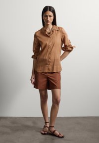 Joven mujer con cabello largo y oscuro que lleva una camisa marrón con botones, pantalones cortos color óxido y sandalias marrones, de pie frente a un fondo claro.