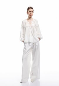 Blusa bianca in cotone con dettagli in broderie anglaise, caratterizzata da scollo a V, maniche lunghe a sbuffo e lacci regolabili. Abbinata a pantaloni bianchi a gamba larga.