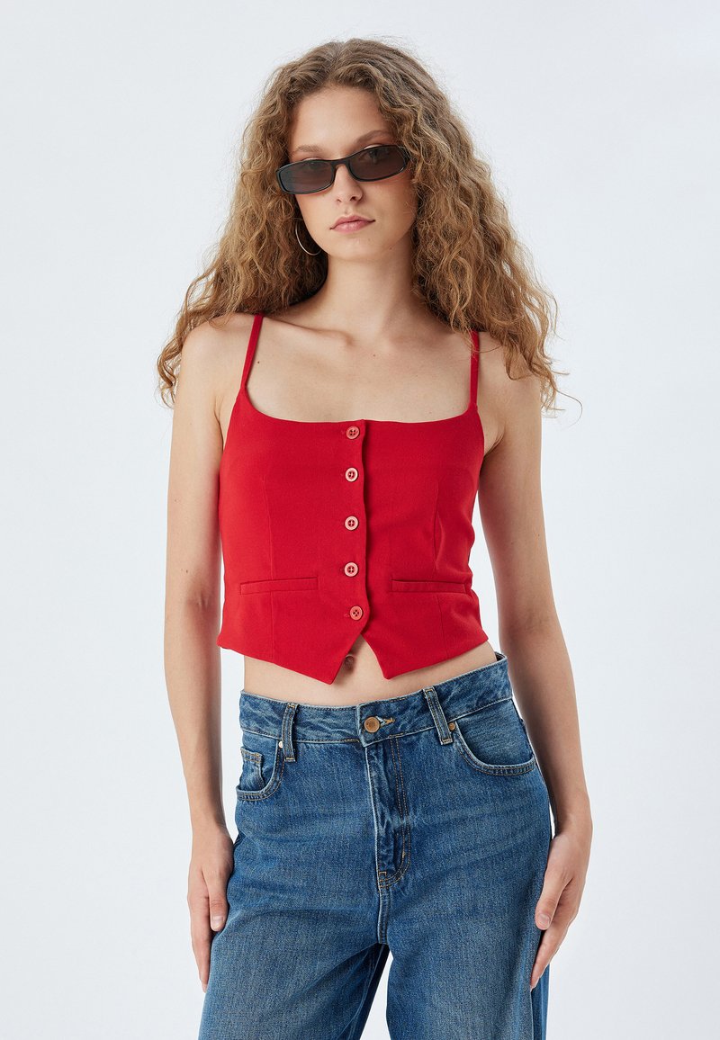 Rotes verkürztes Tanktop mit dünnen Trägern, vorderen Knöpfen und Seitentaschen. Kombiniert mit hochtaillierten, blauen Jeans.